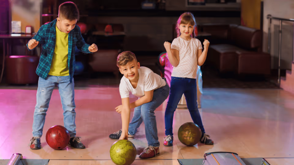 Three children joyfully playing bowling with colorful balls, surrounded by toys like Beanbag Toss and Bowling Sets.
