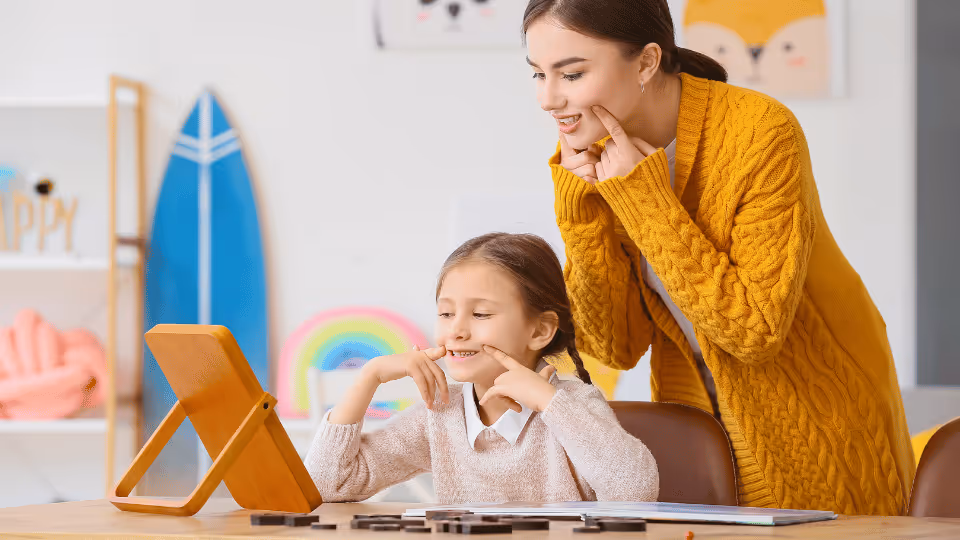 A woman and a child sit at a table, engaging with a tablet, illustrating the theme of differentiating speech and language disorders