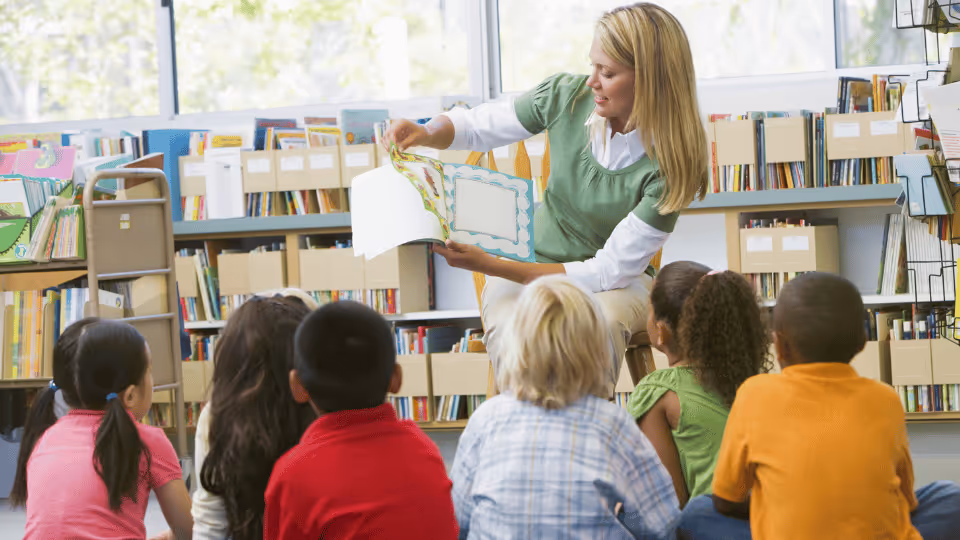 A woman reads to a group of attentive children in a library, surrounded by shelves filled with educational books