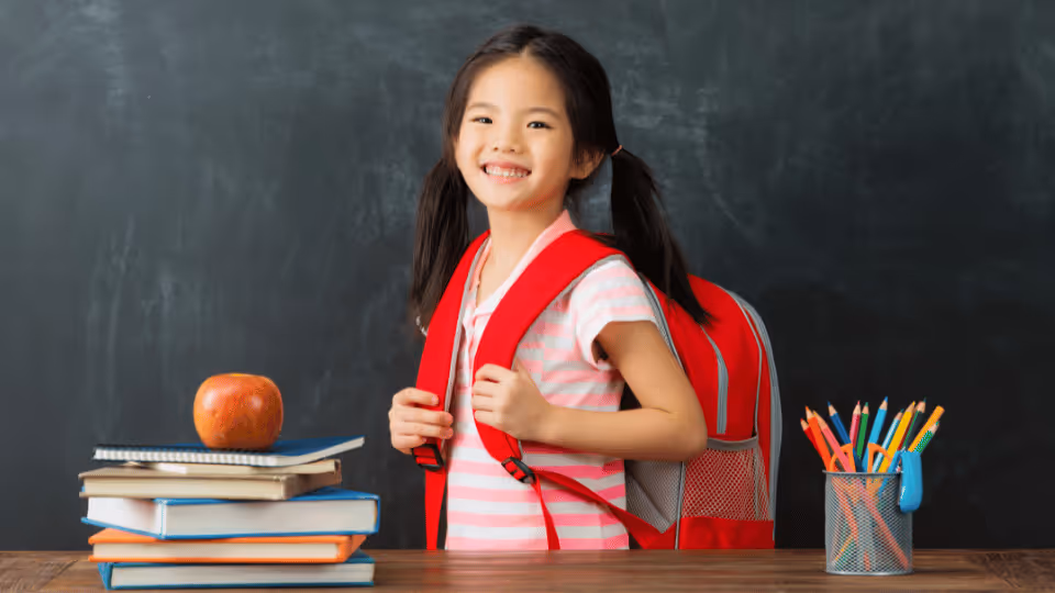 A little girl, equipped with a backpack and books, stands in front of a chalkboard, preparing for her initial treatment session.