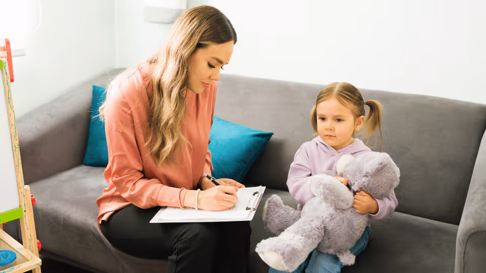 A woman and a young girl holding a teddy bear sit on a couch, symbolizing support in understanding therapy differences.