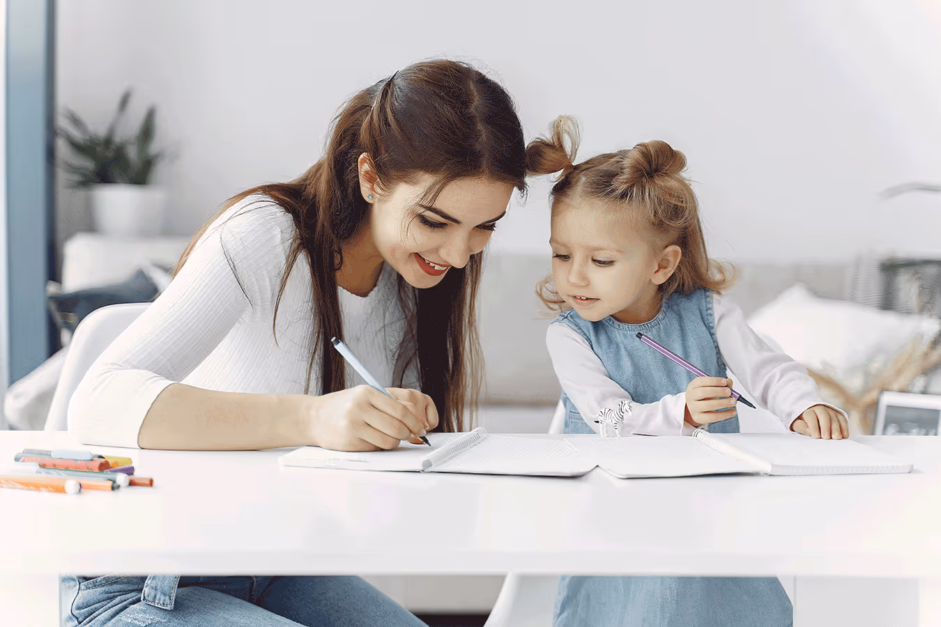 A woman and child engage in handwriting practice at a table, using a notebook and pencils to enhance fine motor skills.