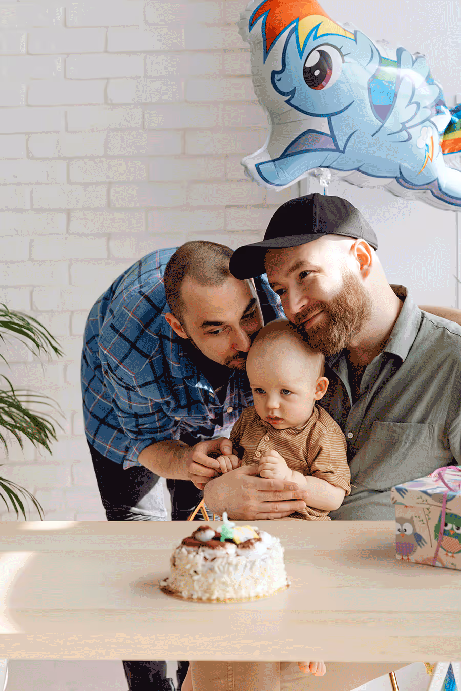 Two dads celebrate a milestone birthday with their baby at a table, featuring a colorful cake.