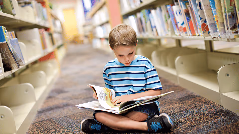 In a Houston public library, a young boy sits on the floor, deeply focused on reading a book amidst the bookshelves.