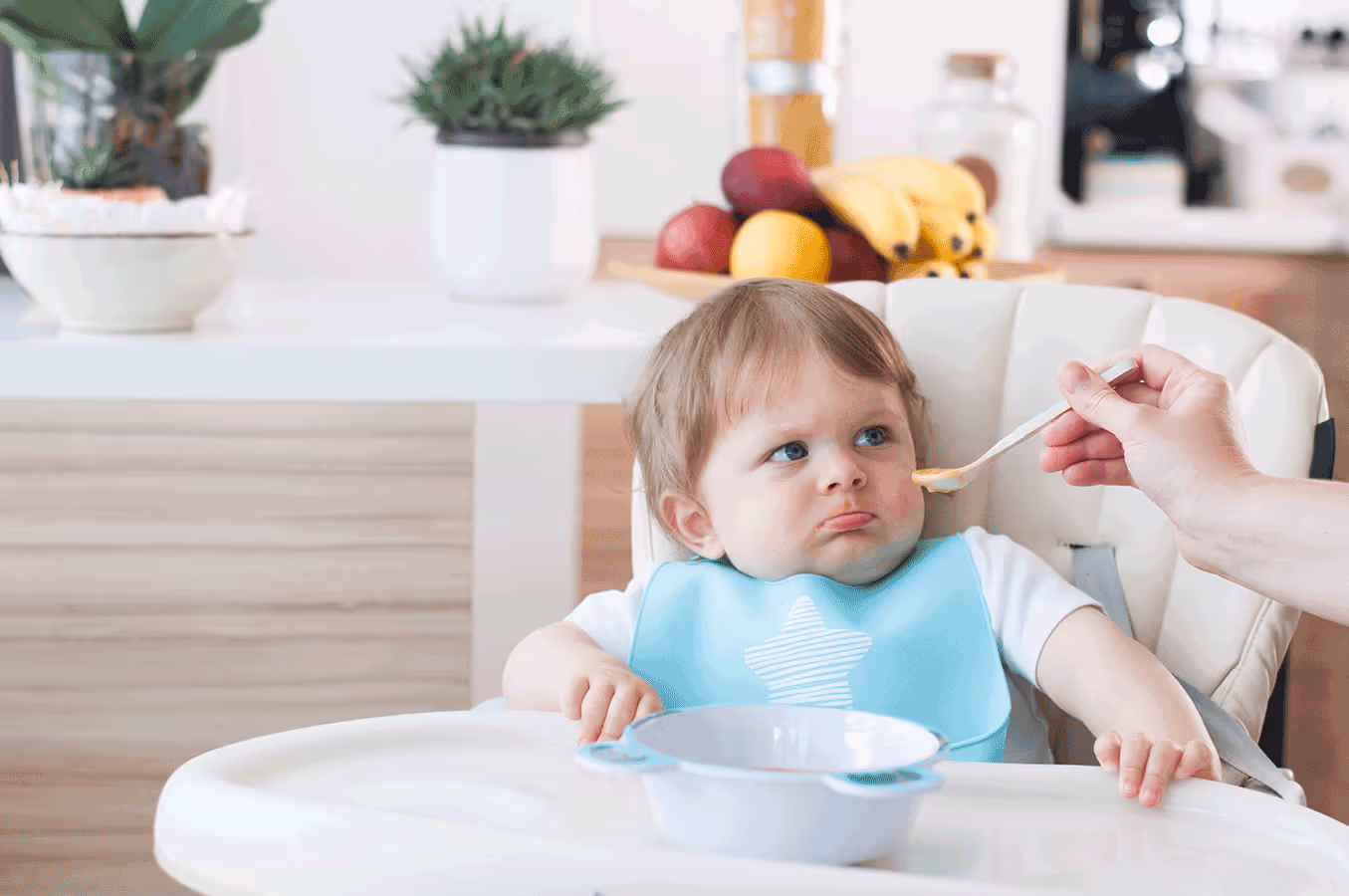 A baby in a high chair with a spoon in his mouth, showing signs of feeding issues during mealtime.