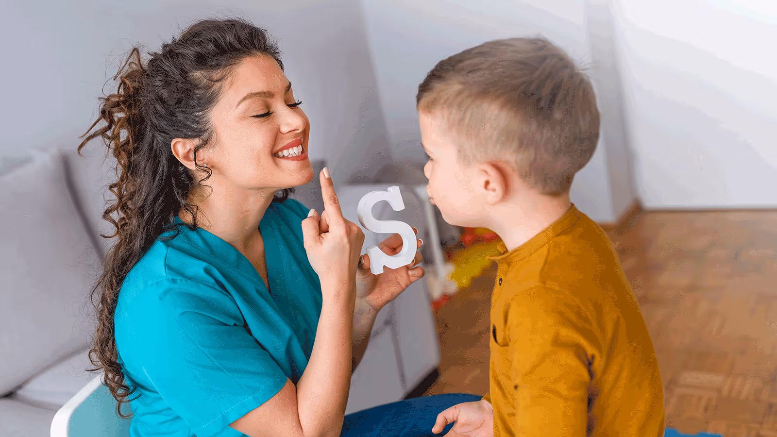 A woman and a child sit on the floor, both smiling while holding up a letter 'S' during a speech therapy session.