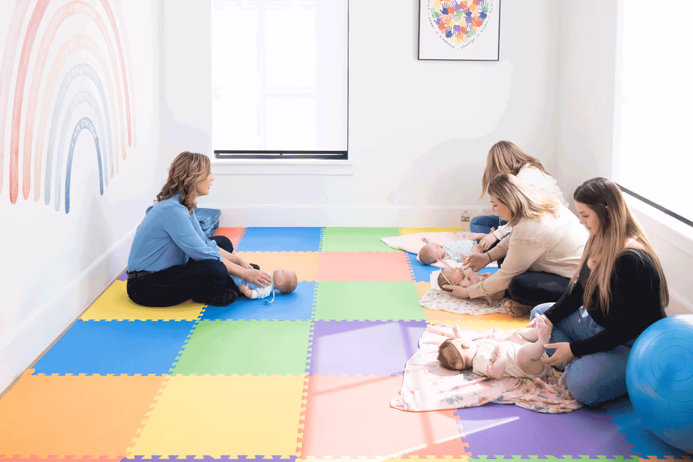 Women and babies enjoy floor time together in a room featuring a vibrant rainbow painted on the wall.