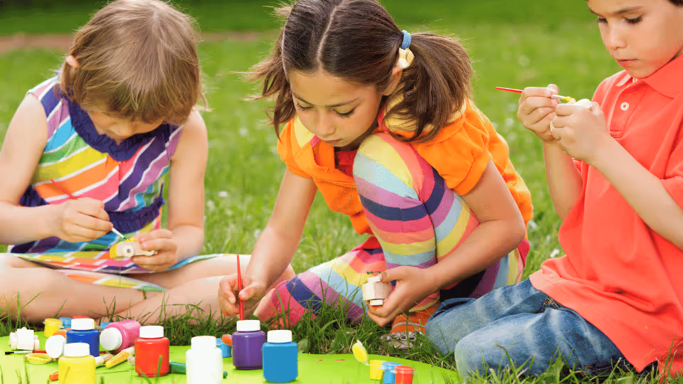 Three children sitting on grass, engaged in outdoor painting activities with brushes, promoting gross motor development.