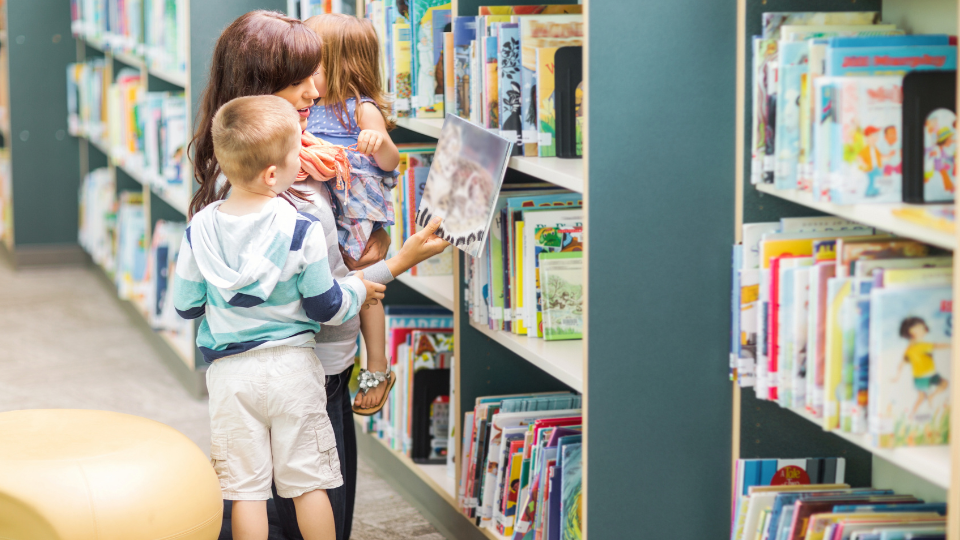A woman and two children browse books together in a Houston public library, surrounded by shelves filled with literature