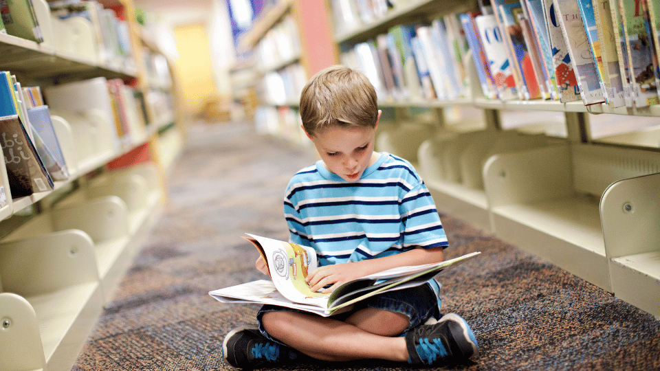 In a Houston public library, a young boy sits on the floor, deeply focused on reading a book amidst the bookshelves.