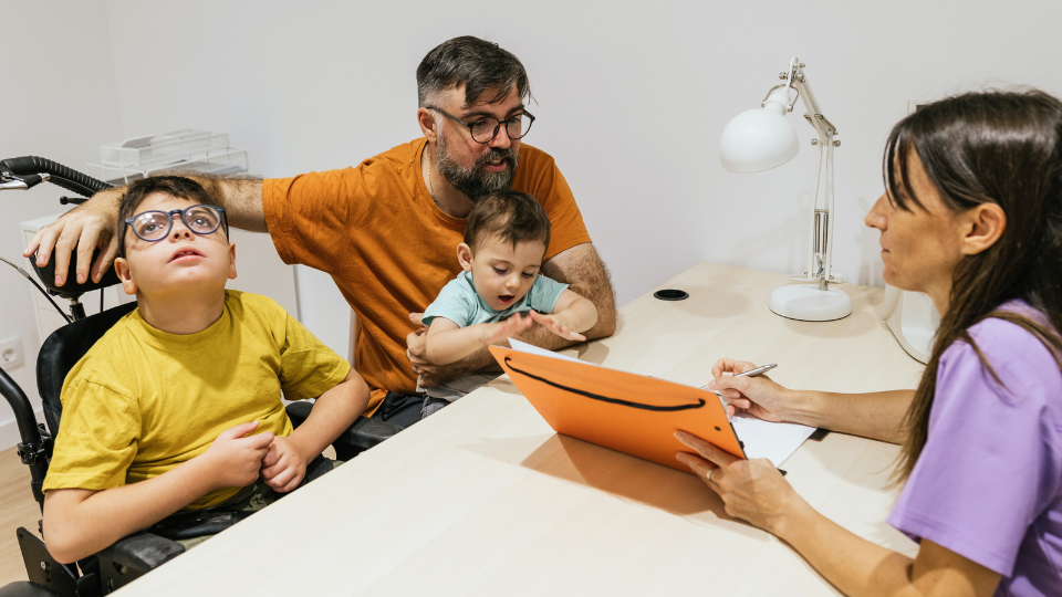 A family gathers around a desk with a man in a wheelchair, engaging in discussions about Boston's developmental disability councils