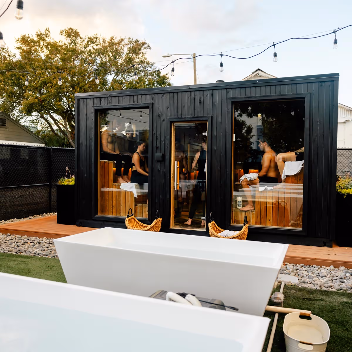 Modern black sauna cabin with large glass windows showing people inside, fronted by white outdoor bathtubs on artificial grass.