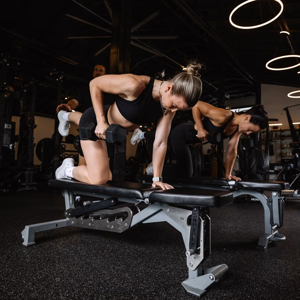 Two women performing dumbbell rows on workout benches in a gym with dark interior lighting.