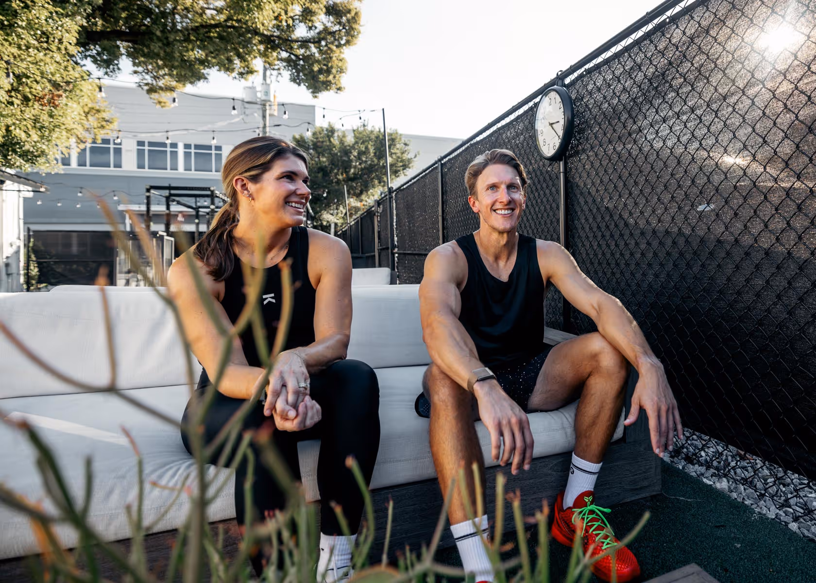 A smiling man and woman in athletic wear sitting on a white outdoor couch near a black chain-link fence with a clock.