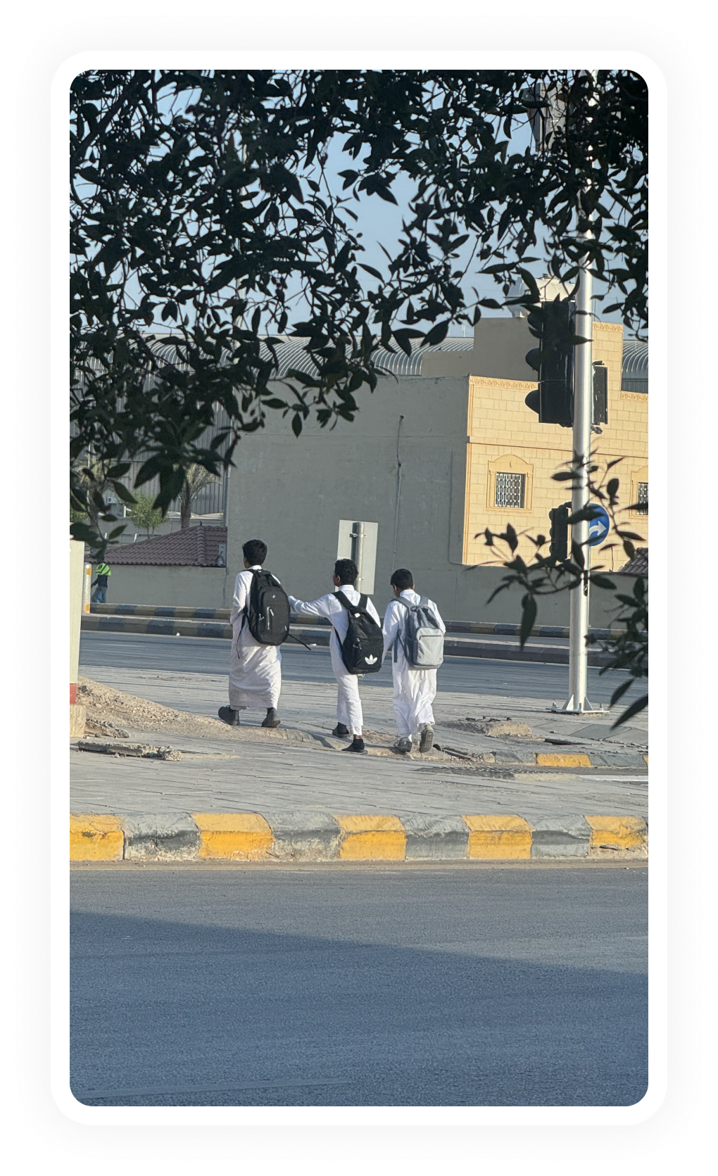 Three boys in white traditional clothes with backpacks walk along a sidewalk near a street.