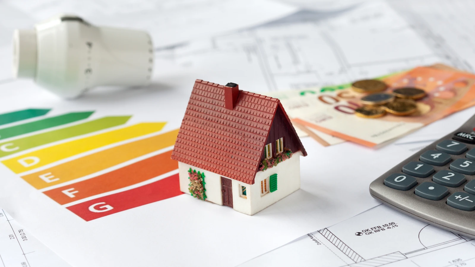 Bild: Miniaturhaus steht auf Sanierungsunterlagen und Thermostat, Taschenrechner und Geld liegen herum