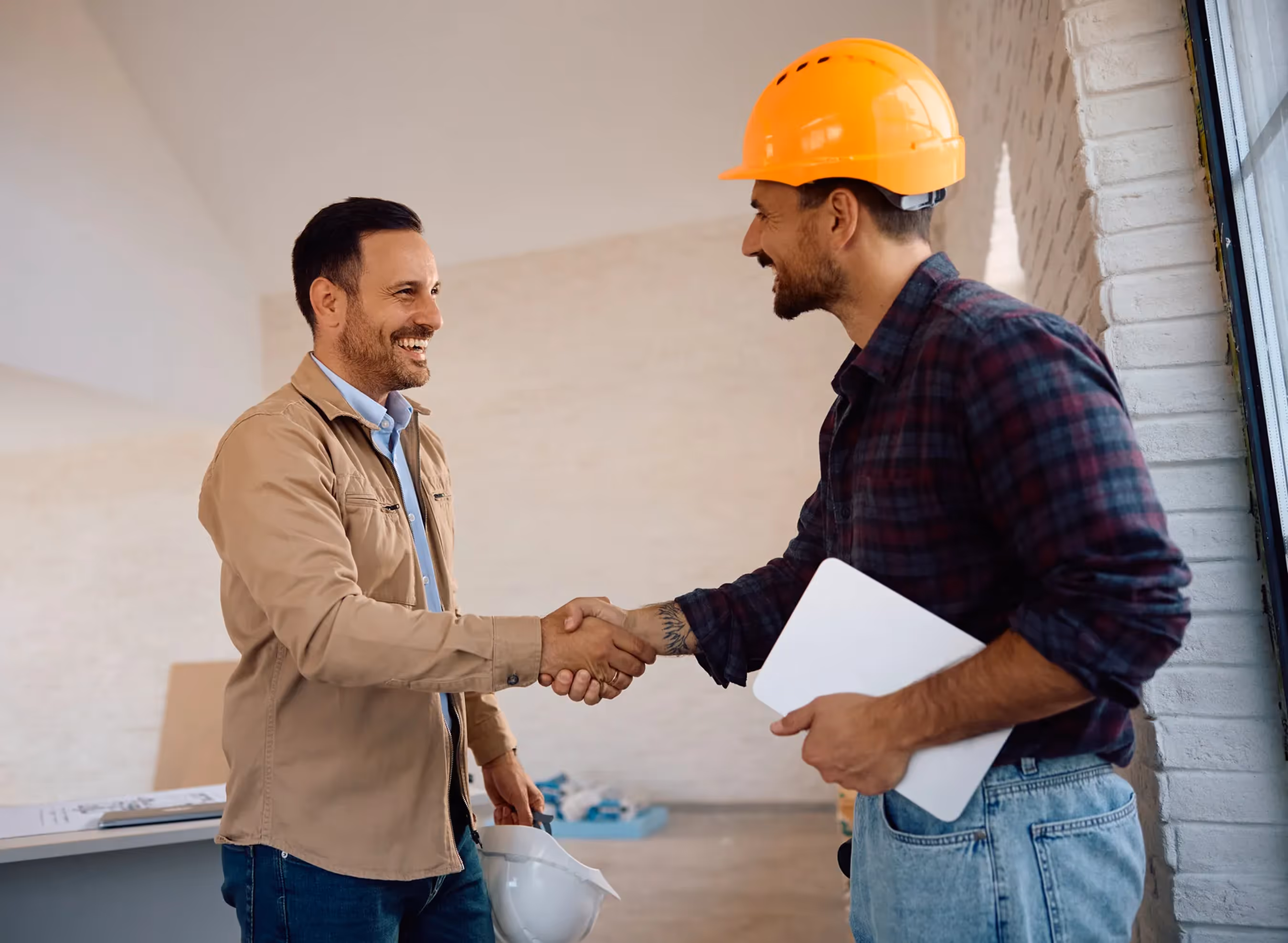 Zwei Männer geben sich auf einer Baustelle die Hand, einer trägt einen Schutzhelm und hält ein Tablet in der Hand.