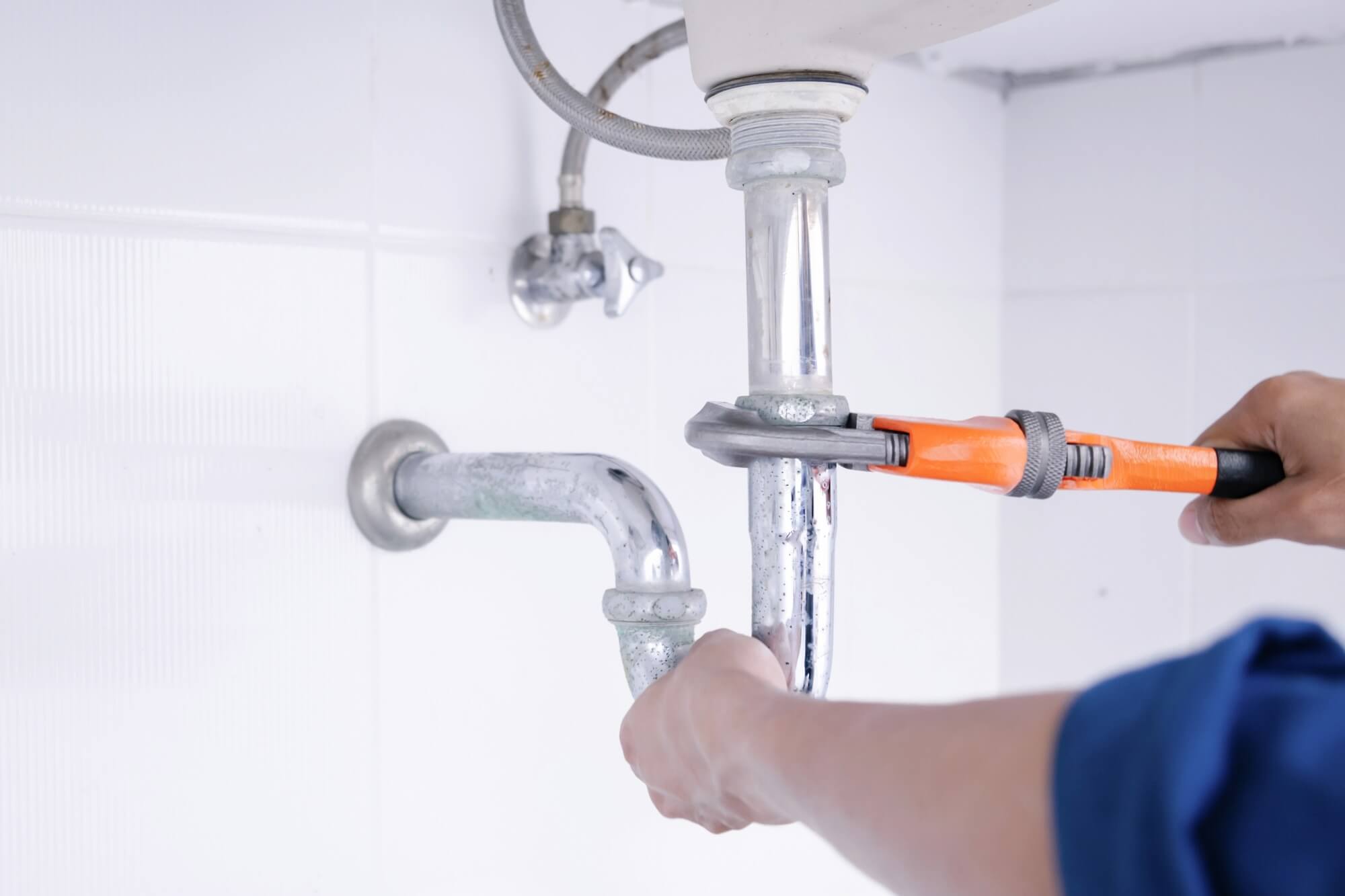 Plumber using a wrench to tighten a chrome pipe under a sink in a bathroom.