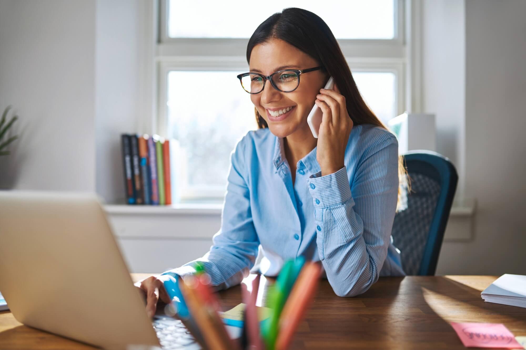 Smiling woman in blue shirt talking on phone while working on a laptop at home office desk.