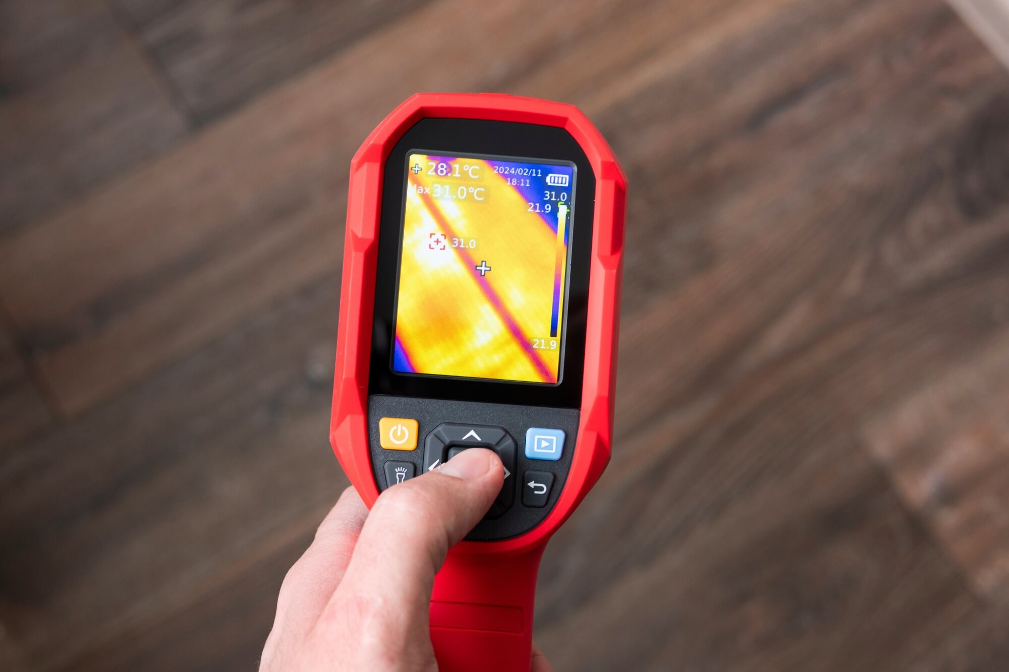 Hand holding a red infrared thermal imaging camera displaying temperature differences on a wooden floor.