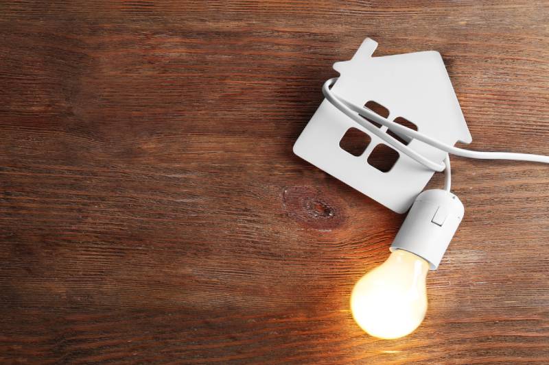 Lit bulb next to house-shaped cutout on wood background.