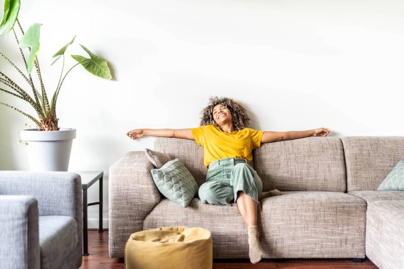 Woman relaxed on couch with arms outstretched, indoor plant nearby.
