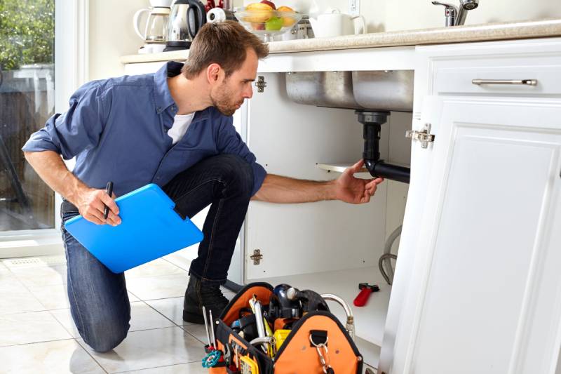 Plumber inspecting kitchen sink pipe with toolkit.
