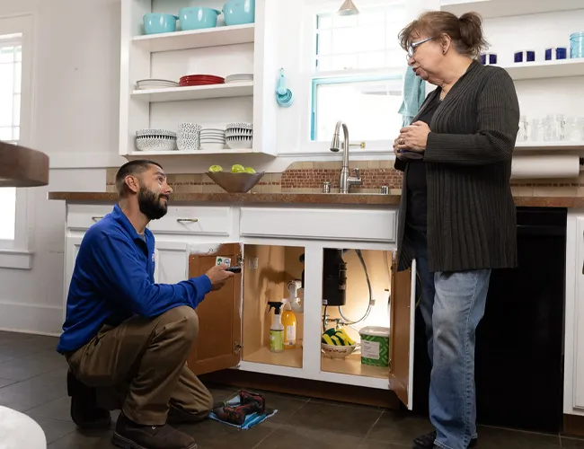Plumber fixing kitchen sink, woman watching.