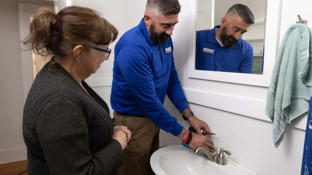 Technician fixing bathroom sink while woman watches.