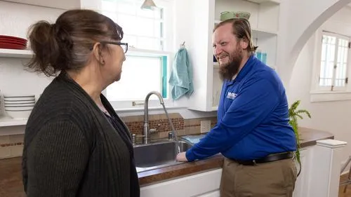 Plumber fixing sink, talking to woman in kitchen.