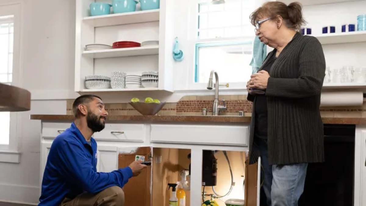 Plumber kneeling under sink talking to concerned woman.