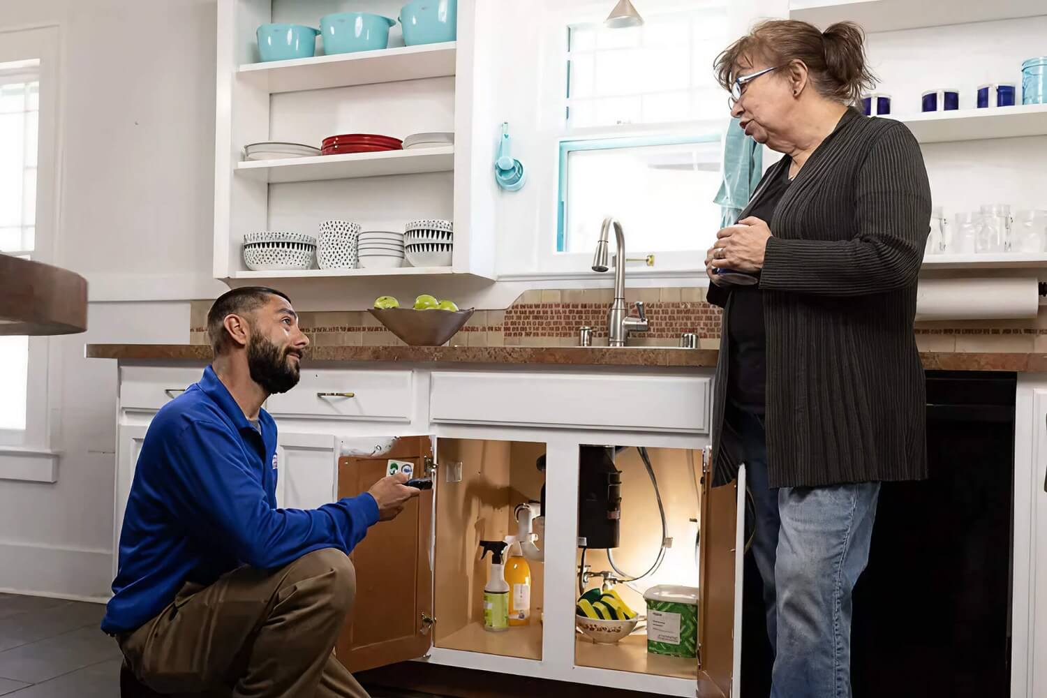 A plumber kneels under a kitchen sink explaining the repair to a homeowner standing nearby.