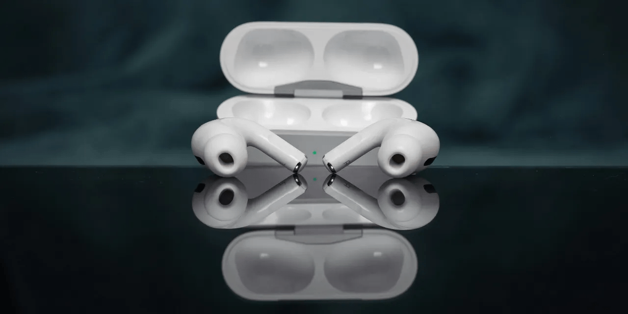 A pair of wireless white earbuds with silicone tips resting on a reflective black surface, positioned in front of an open charging case.