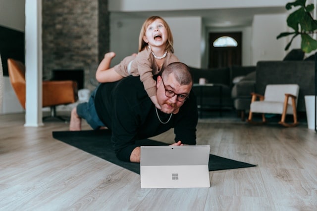 A man planking while looking at his laptop. His daughter is on top of his back smiling.