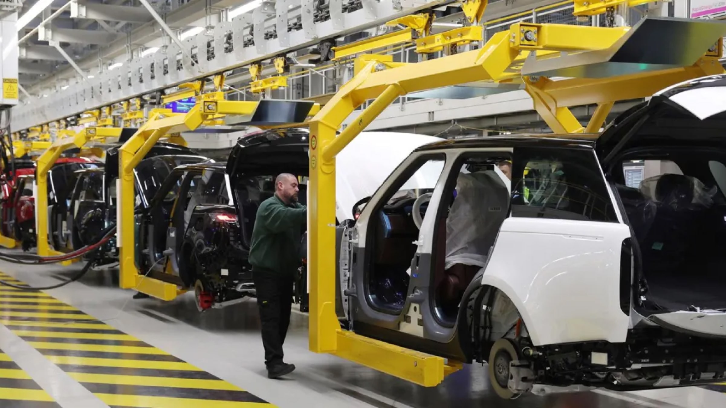 A factory worker inspects vehicles on a Jaguar Land Rover production line, with multiple car bodies suspended by yellow robotic arms inside a modern manufacturing facility.