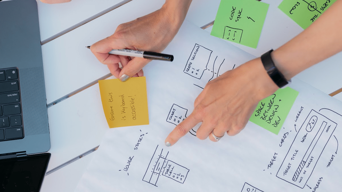 A designer reviews website accessibility sketches and notes, alongside wireframes and accessibility reminders on a desk next to a laptop.