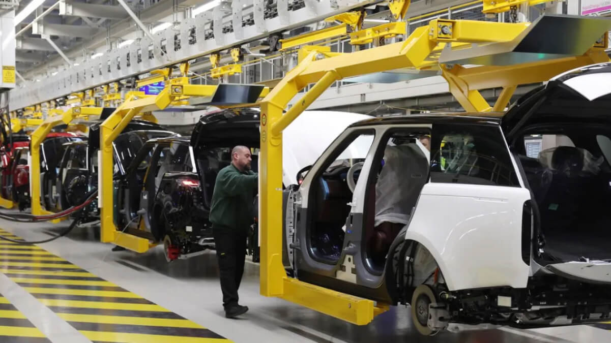 A factory worker inspects vehicles on a Jaguar Land Rover production line, with multiple car bodies suspended by yellow robotic arms inside a modern manufacturing facility.