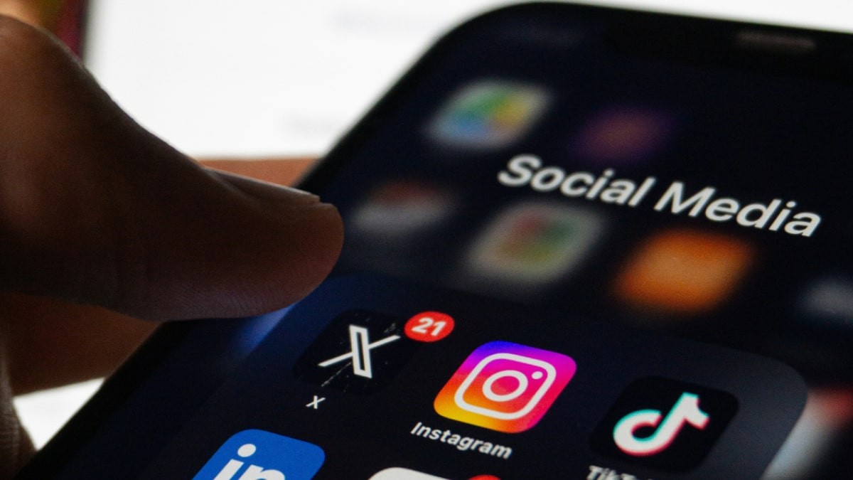 Close-up of a person using a smartphone displaying a folder of social media apps including X, Instagram, and TikTok, under the label “Social Media.”