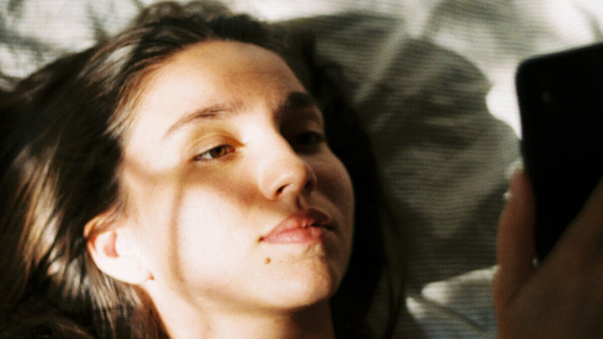 A calm, reflective close-up of a young woman lying in bed, looking at her phone in soft morning light.