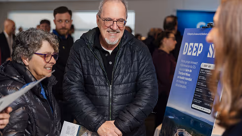 Smiling elderly man and woman wearing glasses and jackets looking at a Deep Sky event poster indoors.