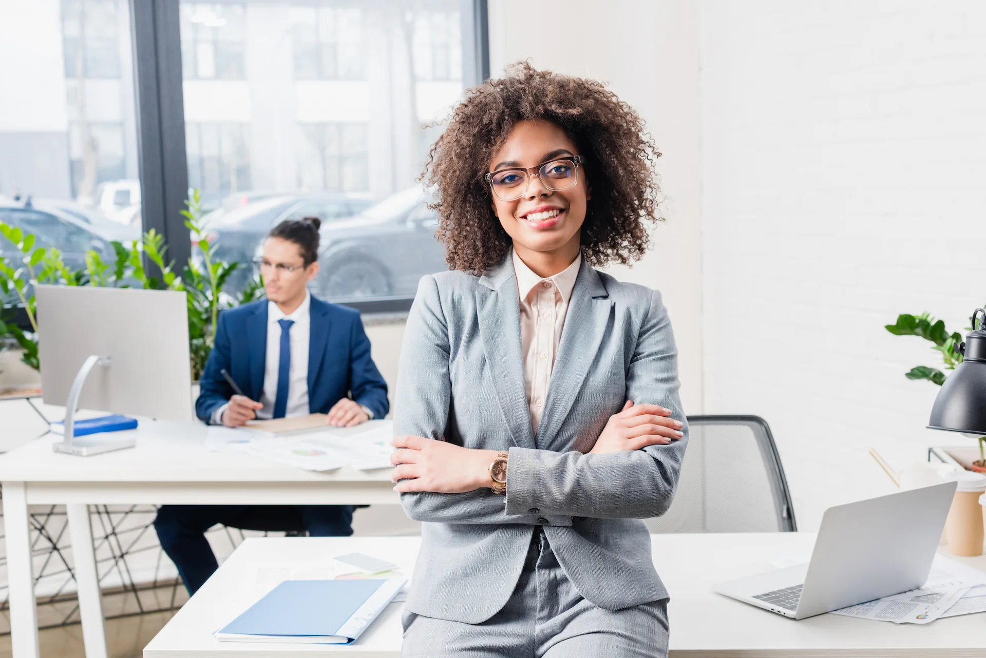 a curly haired woman in a suit smiling while a man works on a laptop in the background