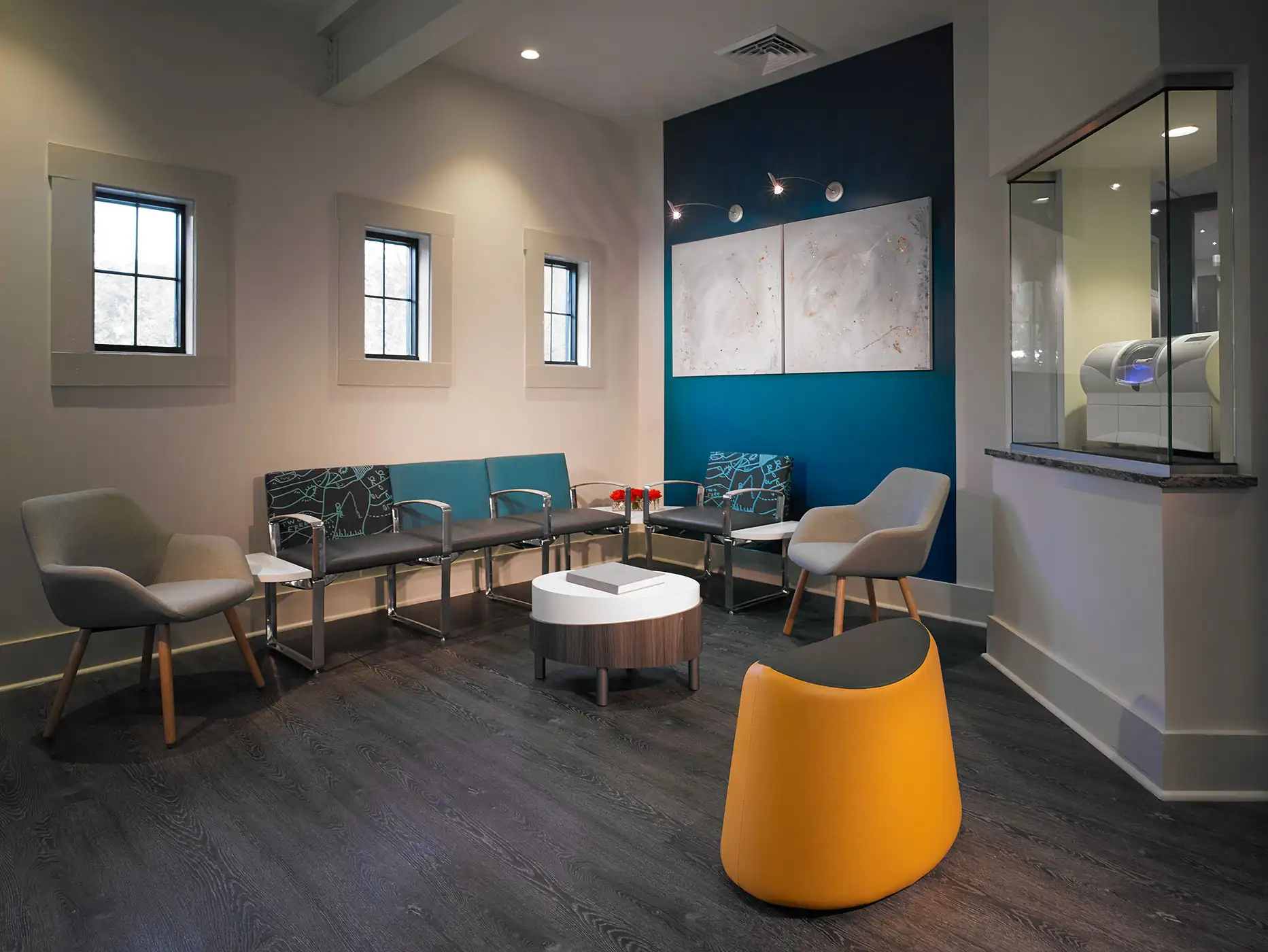 Modern waiting room with gray chairs, teal bench seating, yellow stool, round table, and blue accent wall.