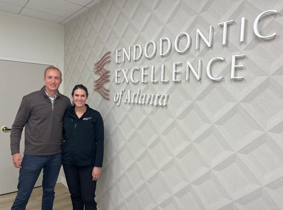 Two smiling people standing in front of a wall sign reading Endodontic Excellence of Atlanta.