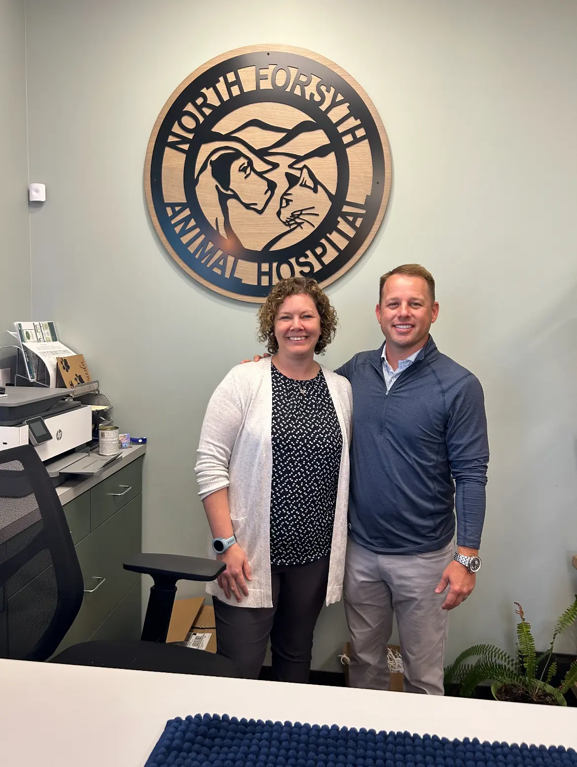 Two smiling people standing in front of North Forsyth Animal Hospital sign inside an office.