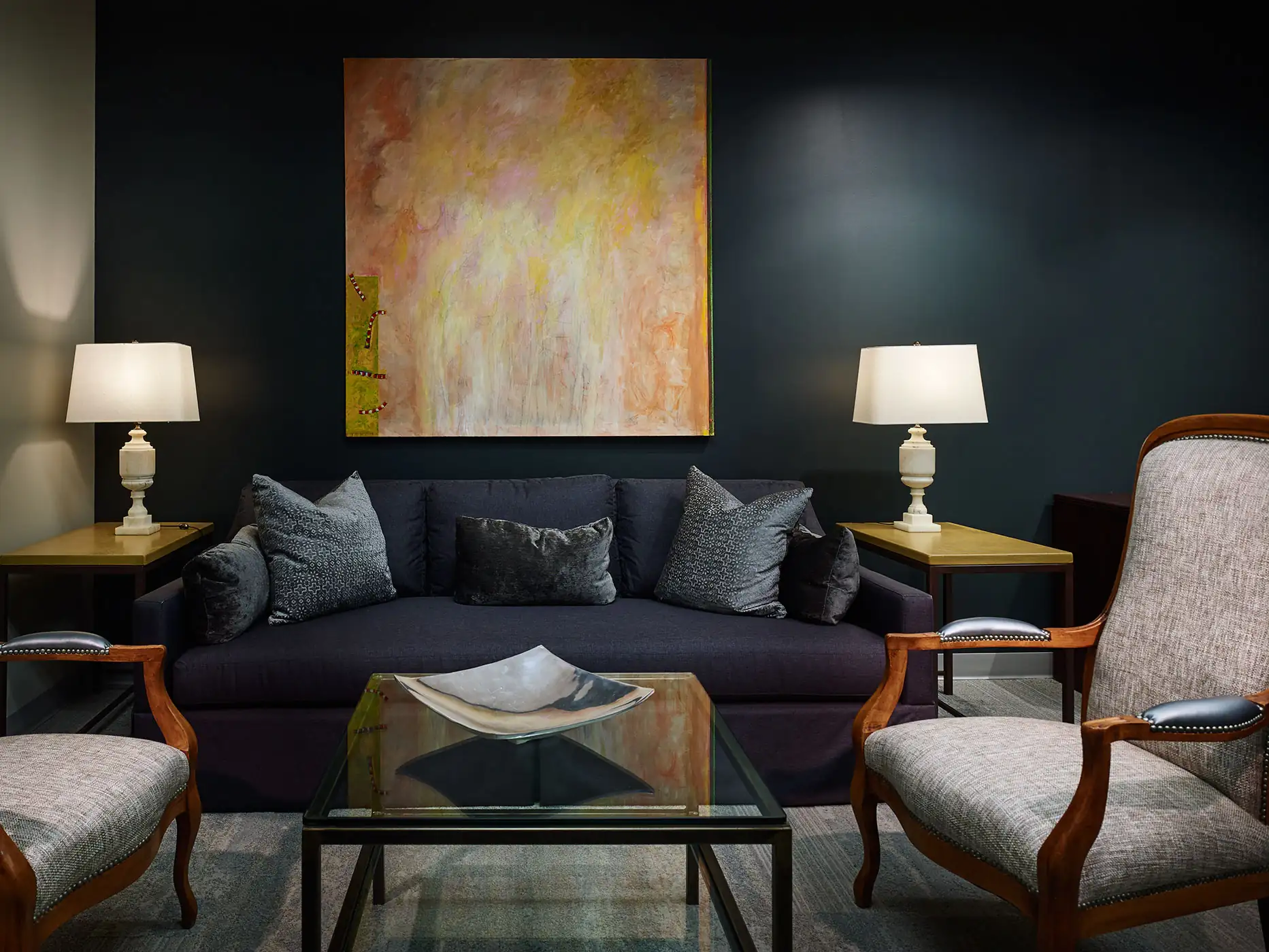 Modern sitting area with dark sofa, patterned cushions, armchairs, glass coffee table, and abstract wall art.