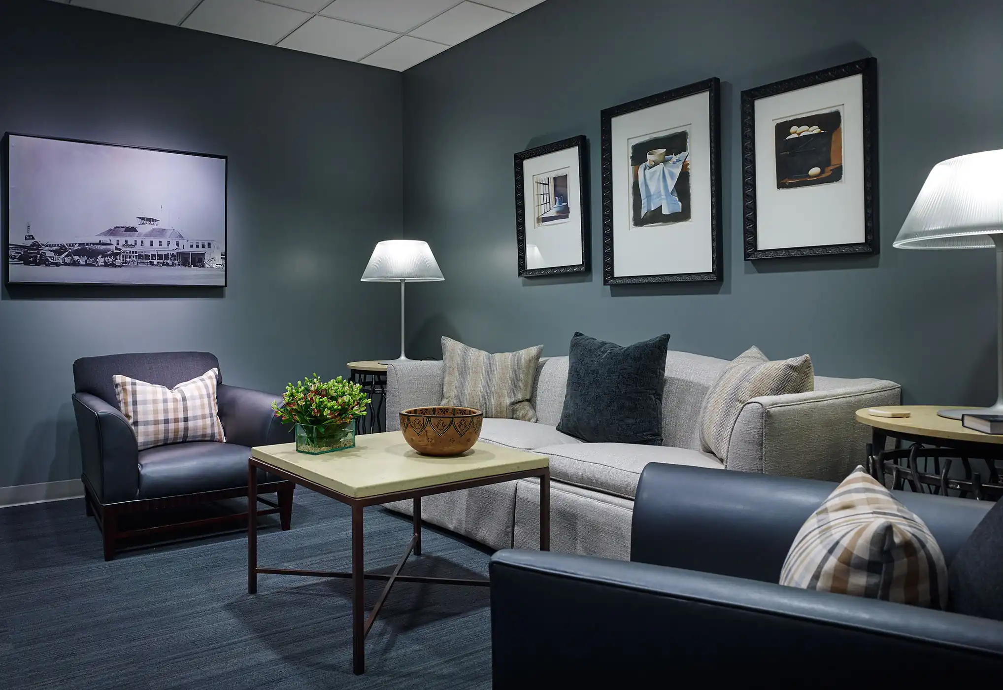 Modern waiting room with gray sofa, armchairs, framed art, lamps, and a central table with flowers and a bowl.