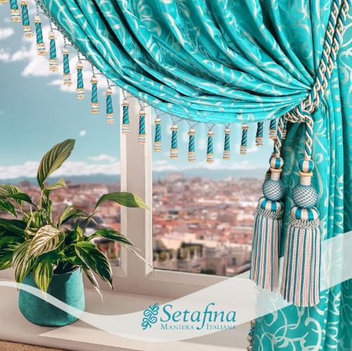 Elegant turquoise curtains with fringes and tiebacks of our Carmen collection adorn a bright window, overlooking a cityscape. A potted plant sits on the windowsill, adding freshness.