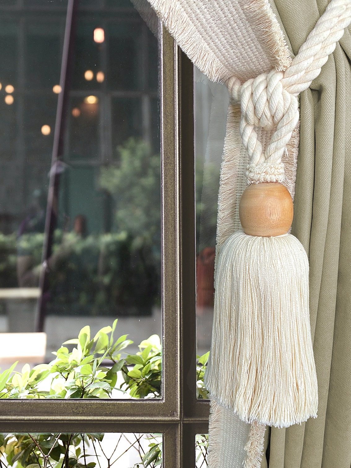 Elegant close-up of a cream-colored curtain tieback from Naturale collection with sunlight streaming through a window, greenery and soft lighting outside. Cozy and serene.