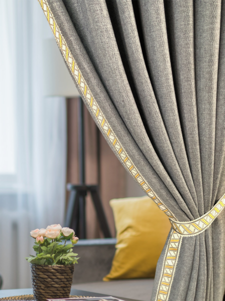 Gray curtain with yellow trims of our Aida Collection embroidery tapes reveals a cozy living room. A yellow pillow and potted flowers add warmth next to a softly lit window.