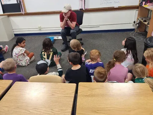 students sitting on a floor while an adult presents a presentation